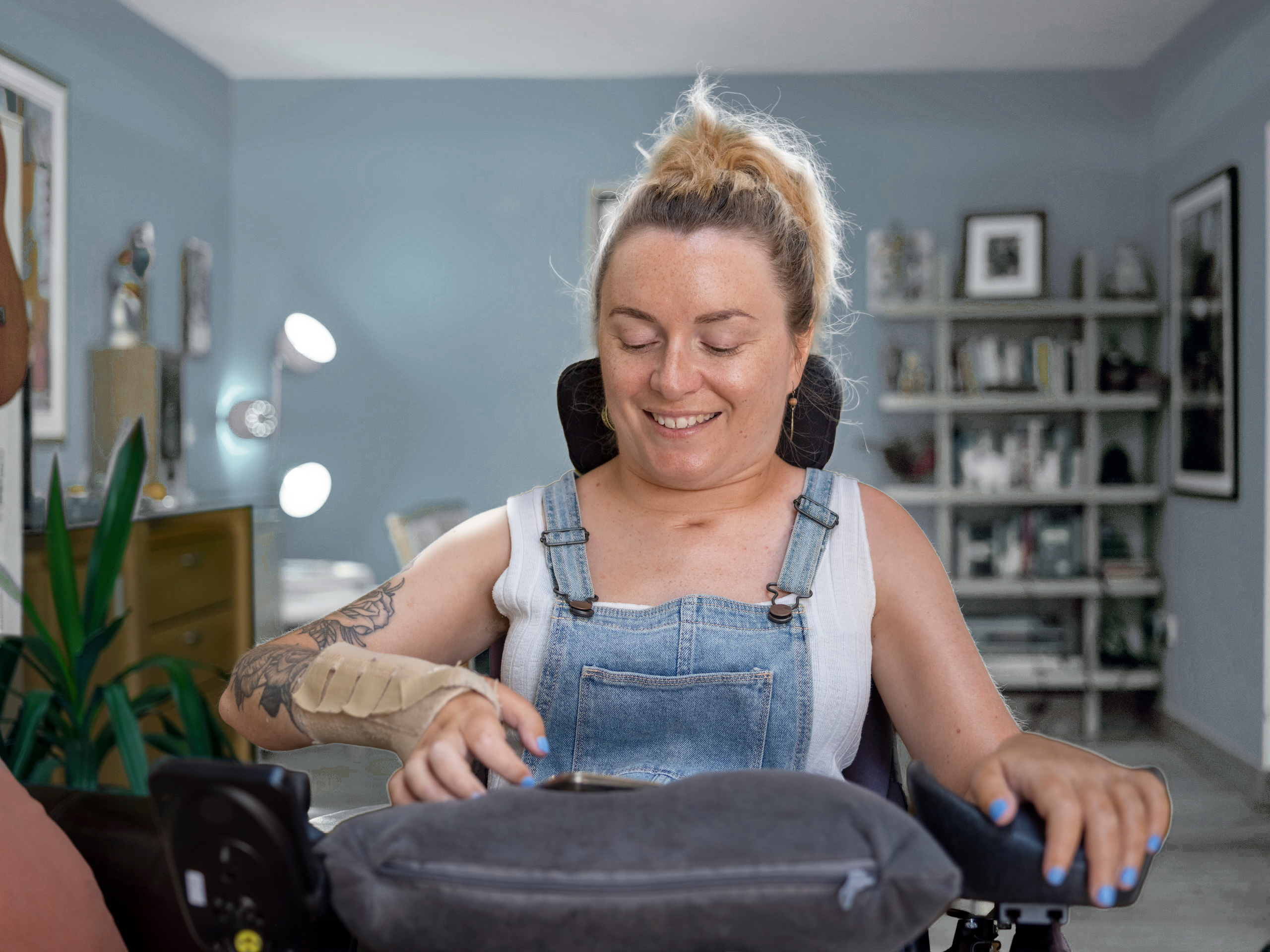 A woman in a wheelchair, wearing denim dungarees and a light shirt, smiles while looking down. She has a plaster on her left wrist and is in a cosy, well-lit room with shelves and plants in the background.