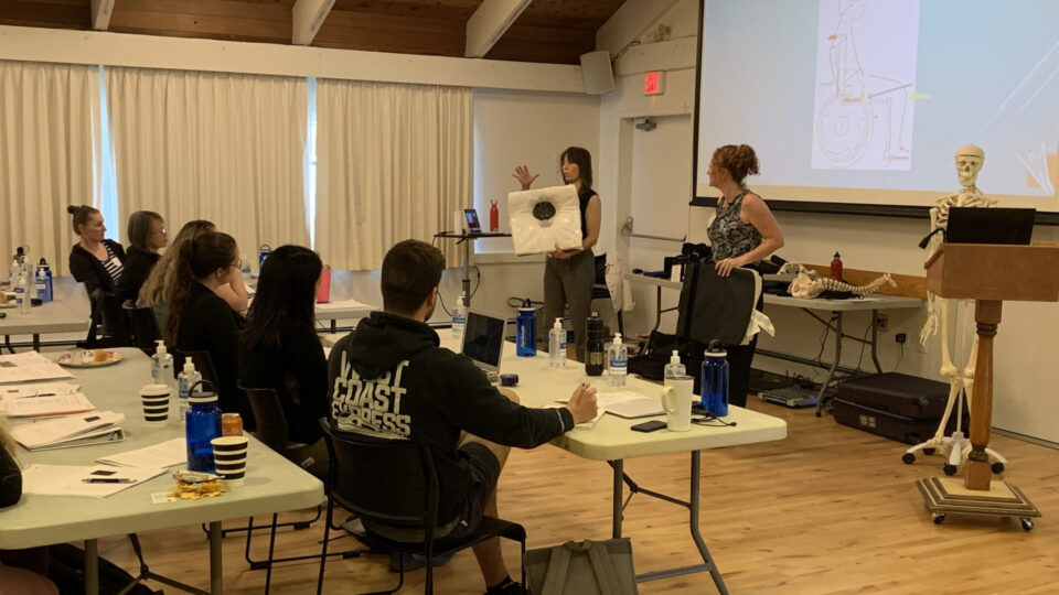A woman stands at the front of a classroom holding up a diagram, while another woman assists. Several people are seated at tables, listening and taking notes. A skeleton model and a projector screen are also visible.